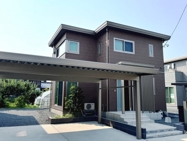 ブラウンを基調とした二階建て住宅の外観です。家の前にはカーポートがあり、広い駐車スペースが確保されています。庭には木や植物があり、緑が映える落ち着いた雰囲気です。