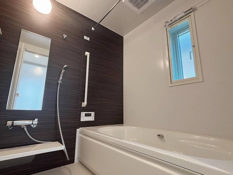 浴槽とシャワーが設置された明るいバスルームの写真です。壁は濃い木目調で、白い手すりと鏡が備えられています。浴槽の隣には小さな窓があり、換気を助けています。天井には換気扇と照明があり、全体的に清潔感のあるデザインです。