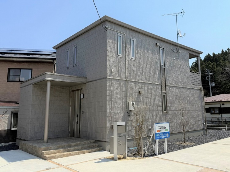 グレーを基調とした二階建て住宅の外観。縦長の窓が特徴で、左側には玄関への庇が設けられています。バルコニーにはアンテナが設置され、敷地内には小さな植栽が並んでいます。