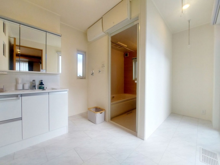 白を基調とした明るい洗面所。左に鏡付きの洗面台があり、右奥にはやわらかな木目の壁に囲まれた浴室が見えます。