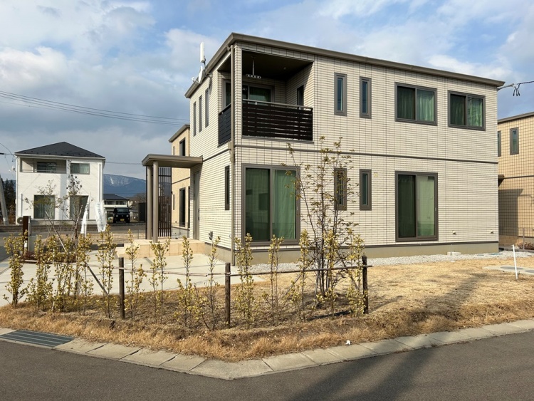 角ばったシルエットが印象的な二階建ての家。ベージュのタイル張りの外壁に、濃い色の窓枠やバルコニーがアクセントになっている。玄関には屋根付きのアプローチがあり、整えられた庭には細い木々が並ぶ。奥には他の住宅や山並みが見え、静かな環境が広がっている。