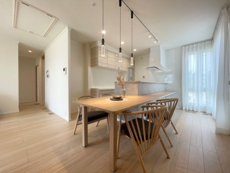 木製の大きなダイニングテーブルとシンプルな椅子が並ぶ明るいダイニングキッチン。モダンなペンダントライトが天井から吊り下がり、窓から柔らかな光が差し込んでいます。