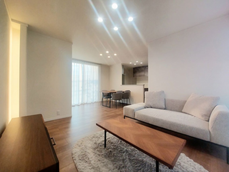 落ち着いた色合いのリビングダイニング。木目のフローリングに、ライトベージュのソファ、コーヒーテーブル、壁際にはローボードと照明付きの装飾コーナーがあり、間接照明で壁が柔らかく照らされている。