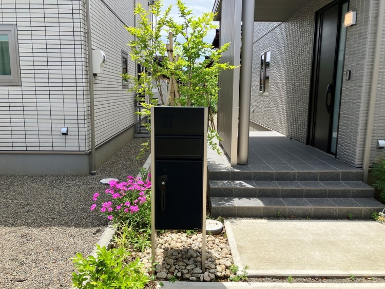 家の玄関前に設置された黒い宅配ボックス。周りには小石と植物が配置されています。