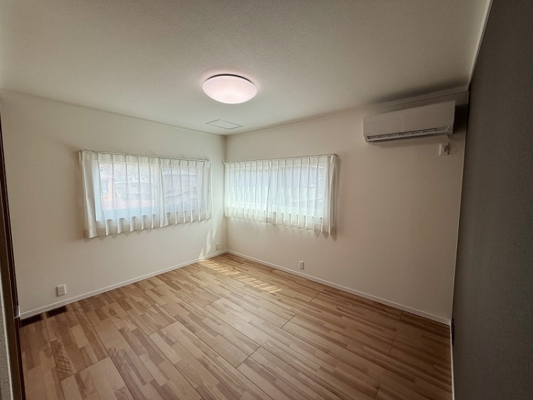 柔らかが風にな光が差し込む角部屋。二面に窓があり、白いレースカーテン揺れる様子が心地よい。木目調の床が温かみを感じさせ、シンプルながらも落ち着いた雰囲気の空間。