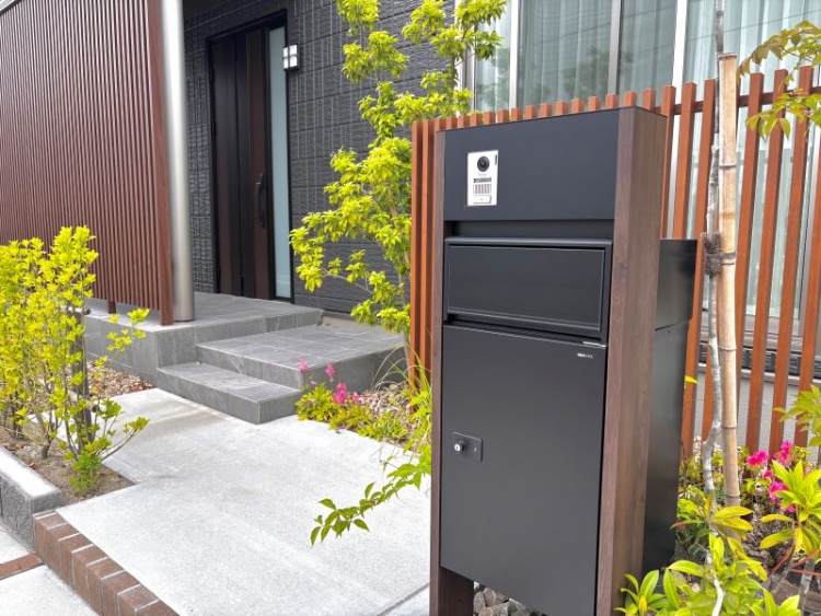 ダークグレーの外壁の２階建て住宅のB棟。玄関前に設置された黒い宅配ボックスと周囲には植栽が植えられています。