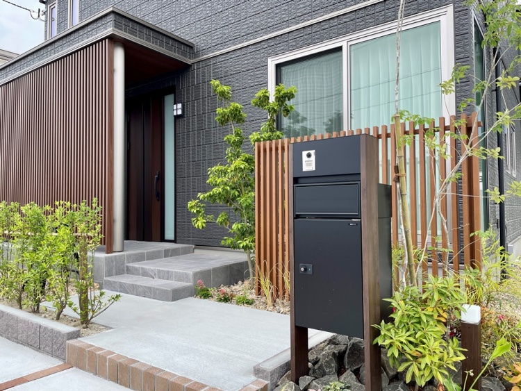 ダークグレーの外壁の２階建て住宅のB棟。玄関前に設置された黒い宅配ボックスと周囲には植栽が植えられています。