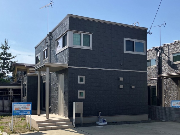 ダークグレーを基調とした二階建て住宅の外観で、シンプルなデザインが特徴です。玄関には小さな階段があり、縦長の窓がスタイリッシュに配置されています。