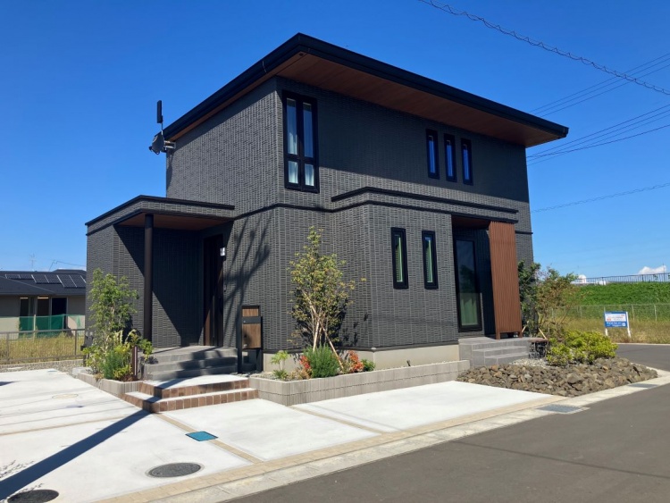 黒い外壁が印象的な二階建ての家で、晴れ渡る青空の下、小さな庭木が生き生きと葉を広げています。