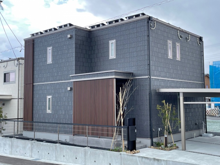 '深みのある青灰色の外壁が印象的な二階建ての家。玄関まわりには縦格子があしらわれ、前には若い木々が植えられています。