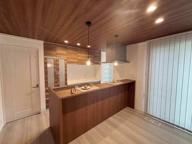 木目調のキャビネットが並ぶキッチンの写真です。中央にはシンプルなカウンターがあり、2つのプレートとカップが並べられ、洋ナシが添えられています。天井からは裸電球のペンダントライトが下がり、カウンターを優しく照らしています。奥には広めの収納スペースがあり、全体的に温かみのあるデザインです。