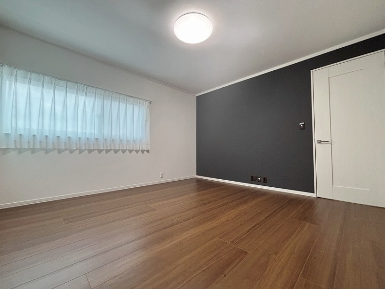 白い壁と木目調の床が特徴のシンプルな洋室です。角にL字型の窓があり、柔らかい光を取り込む白いカーテンが掛かっています。明るく開放的な雰囲気の空間です。