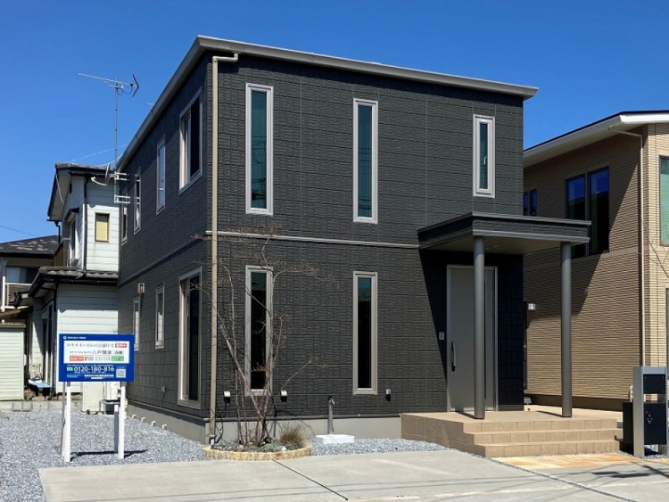ダークグレーを基調とした二階建て住宅の外観で、縦長の窓がいくつも並んでいます。玄関はシルバーのドアが設置され、小さなポーチがあるモダンなデザインです。前庭には若い木が植えられており、整った外観が印象的です。