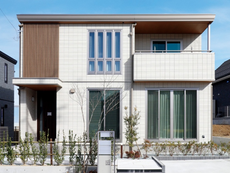 白いタイルの外壁に木目調の格子が特徴的な二階建ての住宅のB棟外観。玄関前には小さな植栽が植えられています。