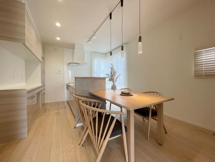 木目調のダイニングテーブルが並ぶ、明るくシンプルなダイニングキッチン。白い壁とペンダントライトが温かみを加え、奥には使いやすそうなキッチンカウンターがあります。