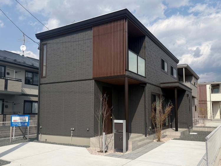 濃いグレーの外壁が特徴的な2階建住宅の外観。縦長の窓と木製の格子がアクセントになっています。前庭には若い木々が植えられており、清潔なコンクリートの駐車スペースがあります。