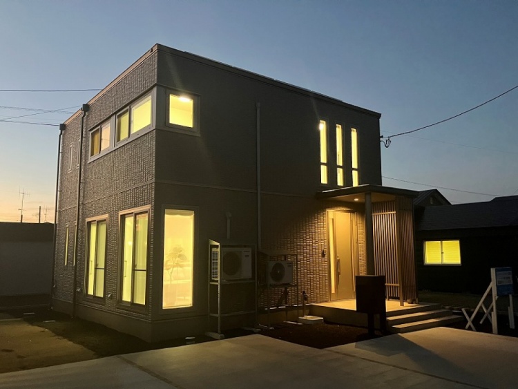 グレーを基調とした二階建て住宅の外観です。縦長の窓と大きなガラス窓が特徴で、玄関にはデザインパネルが設置されています。外壁には2台の空調設備が取り付けられており、玄関には数段の階段が続いています。