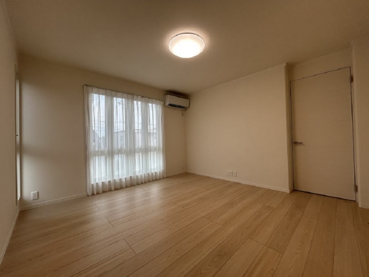 広々とした部屋に、大きな窓から自然光が差し込む穏やかな空間。白いカーテンが優しく揺れ、木目の床が温もりを感じさせる。壁にはエアコンが設置され、快適な居住空間が広がっている。