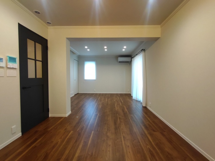 落ち着いた木目調のフローリングが広がるリビング。白い壁と天井が空間を明るくし、大きな窓からは柔らかな光が差し込む。左側には階段があり、奥には収納スペースも備えられている。シンプルながらも温かみのある空間。