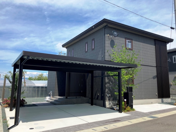 落ち着いた色合いの外壁が特徴的な二階建ての住宅。玄関前には木目調の格子があり、和の雰囲気を感じさせる。カーポートが備えられ、車を停めるスペースも確保。周囲には緑が植えられ、自然の彩りが加えられている。