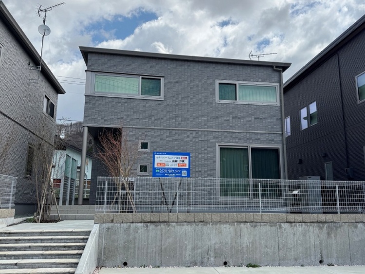 グレーの外壁の２階建て住宅。玄関付近には植栽が植えられています。手前にコンクリートの駐車スペースが見えます。