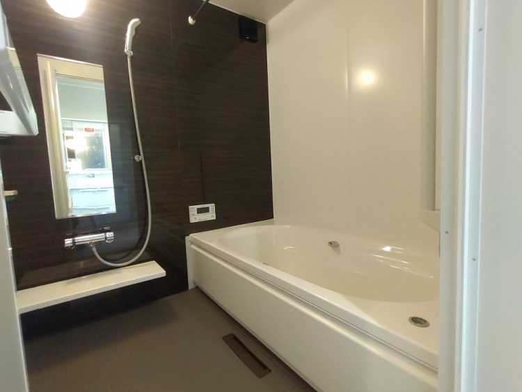 日差しが優しく差し込む広い部屋。木目の床が温かみを感じさせ、白いカーテンが柔らかな雰囲気を演出する。天井には丸い照明があり、エアコンも設置されている。