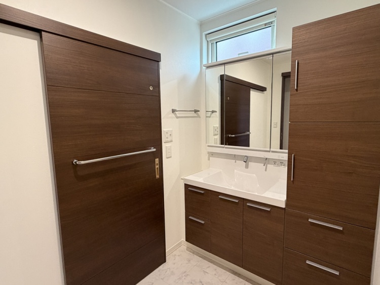 洗面台の上に大きな鏡があり、窓から自然光が差し込んでいます。鏡の前には観葉植物が飾られ、周囲には木目調の収納が配置されています。