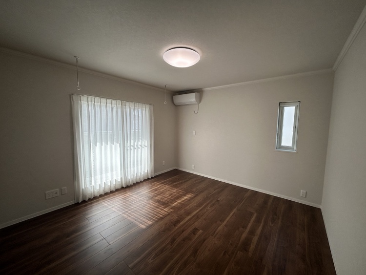 日差しが優しく差し込む広い部屋。木目の床が温かみを感じさせ、白いカーテンが柔らかな雰囲気を演出する。天井には丸い照明があり、エアコンも設置されている。