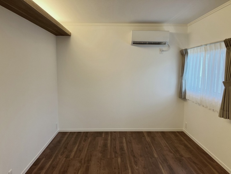 木目調の床が広がる主寝室で、左側に白いドアがあります。天井近くには間接照明が温かみのある光を放ち、右側のエアコンが快適な空間を演出しています。
