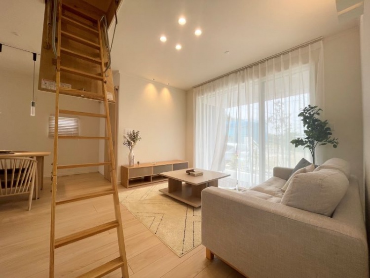 大きな窓から光が差し込むリビングで、白いソファと木のテーブルが並び、天井には小さな階段が伸びています。