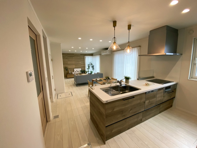 アイランド型のキッチンとリビングが一体化した空間で、キッチンカウンター越しに広がるリビングには大きな窓から自然光が差し込みます。木目調の壁と床が温かみを加え、シンプルな家具と観葉植物が落ち着いた雰囲気を演出しています。吊り下げられたペンダントライトが、スタイリッシュなアクセントとなっています。