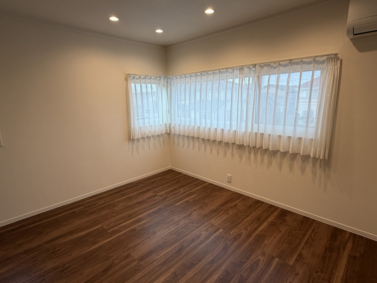 木目調の床と白い壁が特徴のシンプルな主寝室で、天井にはダウンライトが配置されています。大きな窓には白いカーテンが掛けられ、壁にはエアコンが取り付けられています。奥にはウォークインクローゼットが見えます。