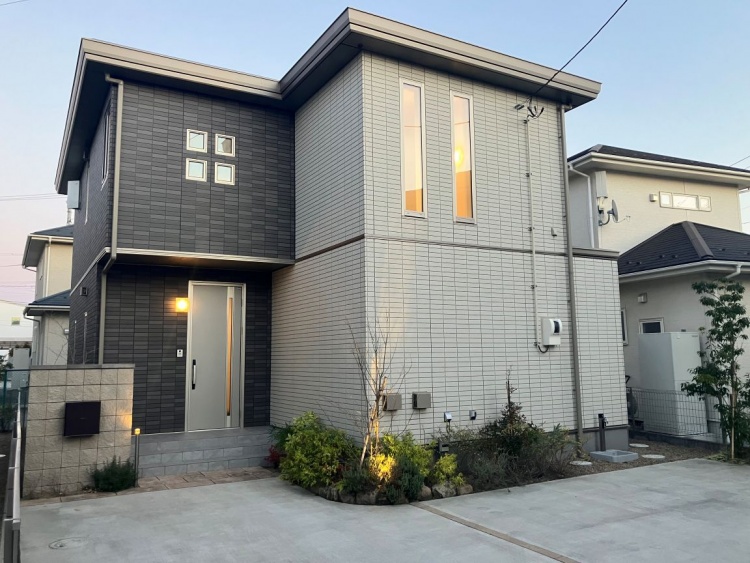 ツートンカラーの現代的な二階建て住宅。左側はダークグレー、右側はライトグレーの外壁で構成され、スリムな縦長窓が特徴的です。