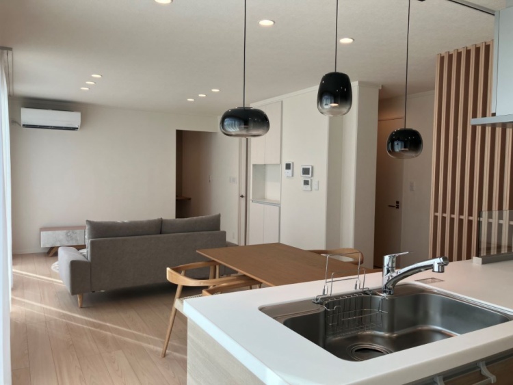 木目調のキッチンと白を基調とした室内が調和し、奥にはソファが置かれた落ち着いたリビングが広がっています。