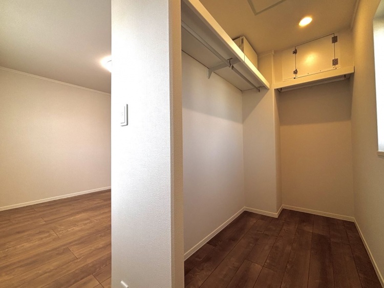 木目調の床に白い壁の主寝室ウォークインクローゼット。上部には収納棚があり、洋服を掛けられるバーが設置されています。奥には明かりが灯り、明るい空間です。