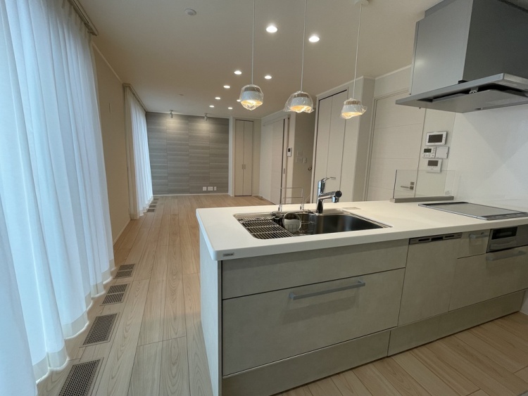 手前に広い調理台があり、その奥に木の床の居間が続いています。天井の灯りと窓の光が重なり、料理をしながら家の気配を感じられる空間です。