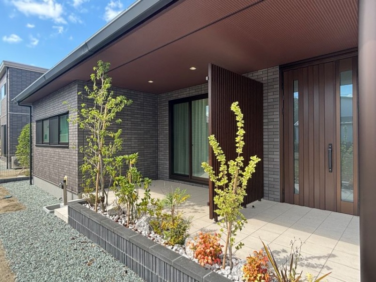 シンプルモダンな外観に、木目調の玄関ドアと縦格子がアクセントを加えるエントランス。整えられた植栽とタイル張りのアプローチが、上質な住まいの印象を演出しています。