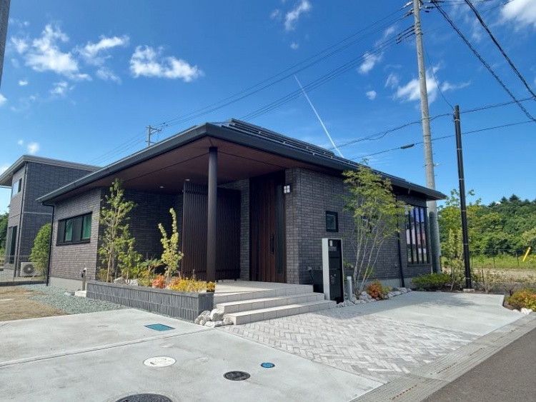 スタイリッシュな平屋住宅の外観。ダークグレーのタイル外壁と深い軒が重厚感を演出し、アプローチには石畳風の舗装が施されており、高級感ある佇まいが魅力的です。