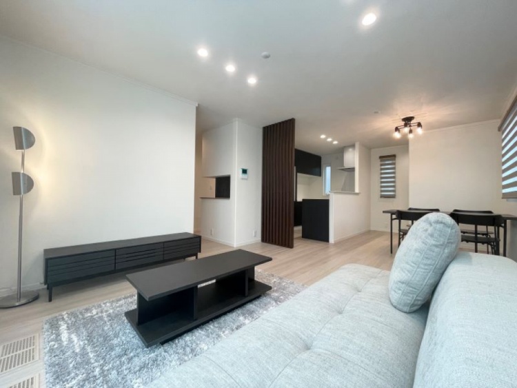 明るいリビングに淡い色のソファと黒いテーブルが置かれ、奥には食卓とキッチンが見えています。