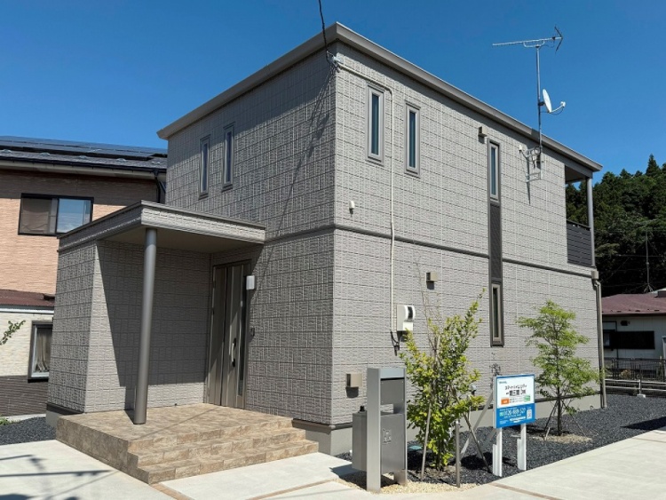 グレーを基調とした二階建て住宅の外観。縦長の窓が特徴で、左側には玄関への庇が設けられています。バルコニーにはアンテナが設置され、敷地内には小さな植栽が並んでいます。