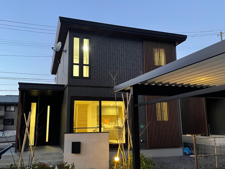 スタイリッシュな外観が際立つ夕暮れ時のモダン住宅。木目調ルーバーとブラックの外壁が高級感を演出し、室内のあたたかな灯りが帰宅を迎えてくれます。