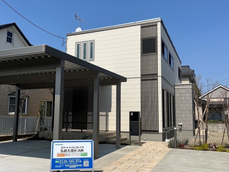 明るいベージュの外壁に黒い縦型のデザインパネルがアクセントとなっている二階建て住宅。前にはカーポートと広い駐車スペースがあり、植栽が家の周りを彩っています。青空の下、清潔感とスタイリッシュさが際立つ外観です。