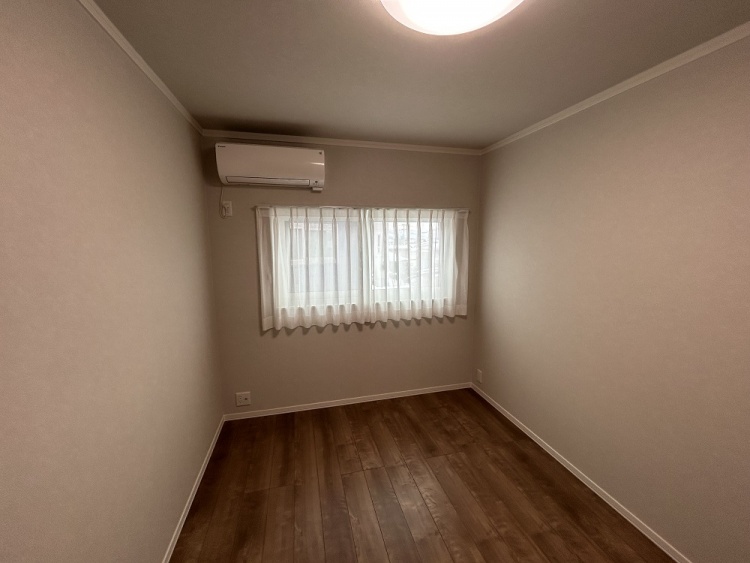落ち着いた雰囲気の部屋。木目調の床に、ベージュの壁紙が温かみを感じさせる。窓には白いカーテンがかかり、エアコンが設置されている。優しい照明が部屋を照らしている。