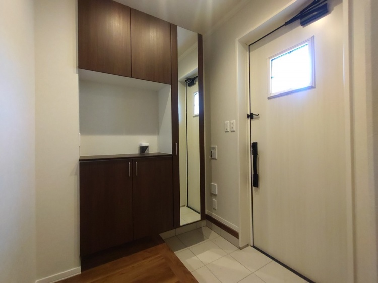 玄関には白いドアが設置され、小さな窓から優しい光が差し込んでいる。壁際には木目調のシューズボックスと姿見が備えられ、機能的で温かみのある空間を演出している。床はタイル張りで、清潔感のある印象を与えている。