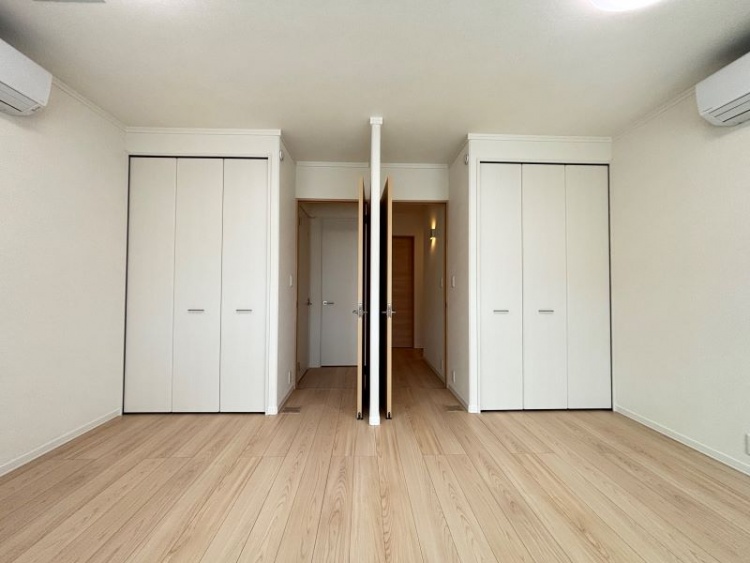 広々とした明るい洋室。白い壁と木目調のドア、エアコンが設置され、窓からは柔らかな光が差し込んでいます。床はナチュラルな木目調のフローリングです。