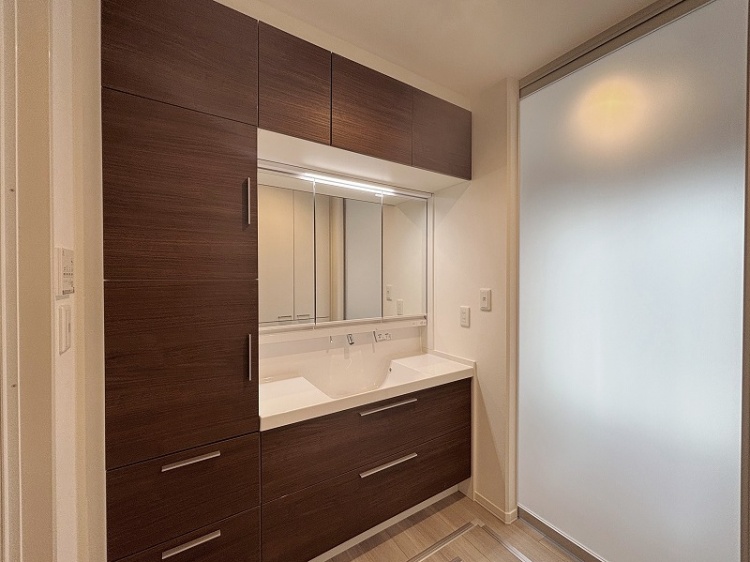 落ち着いた木目調の洗面台がある清潔な空間。大きな鏡が備え付けられ、収納も豊富で使い勝手が良さそう。奥にはバーチカルブラインドがかかった窓があり、柔らかな光が入り込む。