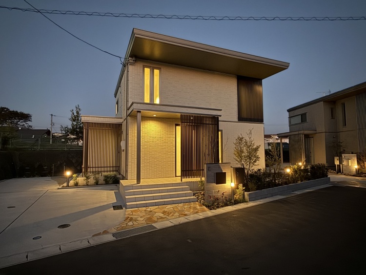白い外壁が特徴的な2階建て住宅の外観。シンプルで直線的なデザインが特徴的で、玄関横には縦格子の目隠しが設置されています。手前には階段と植栽があり、外観は整然として洗練されています。