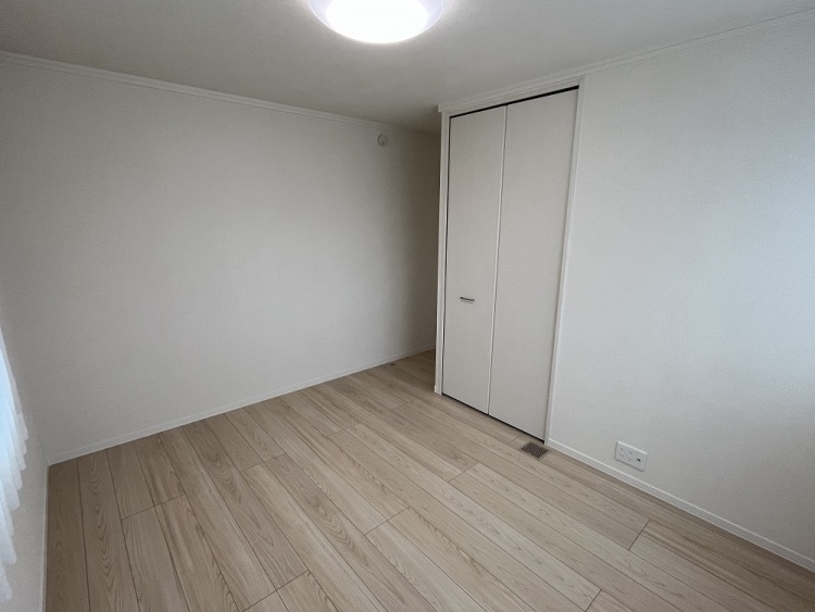 白い壁と木目の床が広がるシンプルな部屋。奥にはクローゼットの扉があり、左側の窓から優しい光が差し込んでいる。温かみのある落ち着いた空間。