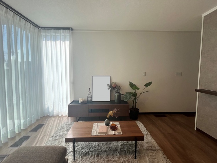 明るい窓際のリビングルームで、落ち着いた木製家具と観葉植物が置かれています。テーブルの上には小物が並び、窓からの光が部屋全体に広がり、リラックスできる雰囲気を演出しています。