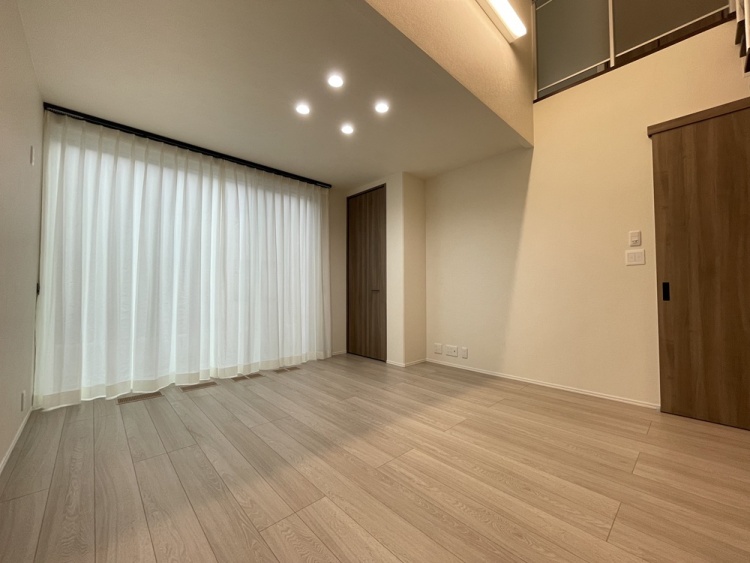 吹き抜け下に広がるリビング空間です。大きな掃き出し窓にレースカーテンが設置され、ダウンライトで明るく照らされています。